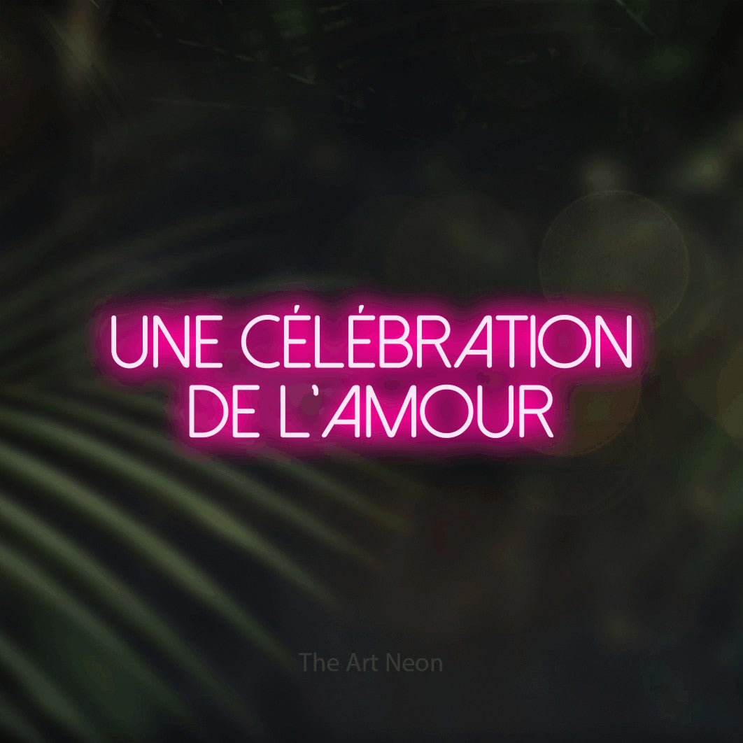 “Une célébration de l'amour” LED neon sign in hot pink, with thin sans-serif lettering across 2 lines, shown against a blurred palm and bokeh-light backdrop.