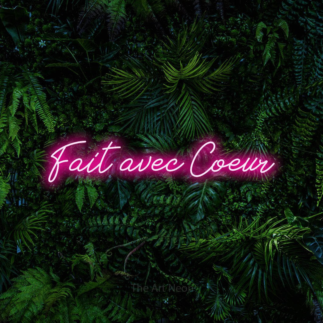 “Fait avec cœur” LED neon sign in hot pink, with cursive lettering, shown against dense tropical foliage.