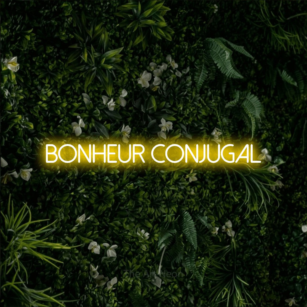 “Bonheur conjugal” LED neon sign in yellow, with thin sans-serif lettering, shown against a dense green plant wall.