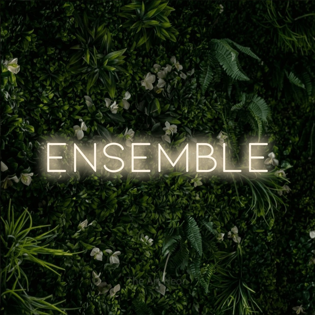 “Ensemble” LED neon sign in warm white, with thin, widely spaced lettering, shown against a dense green plant wall.