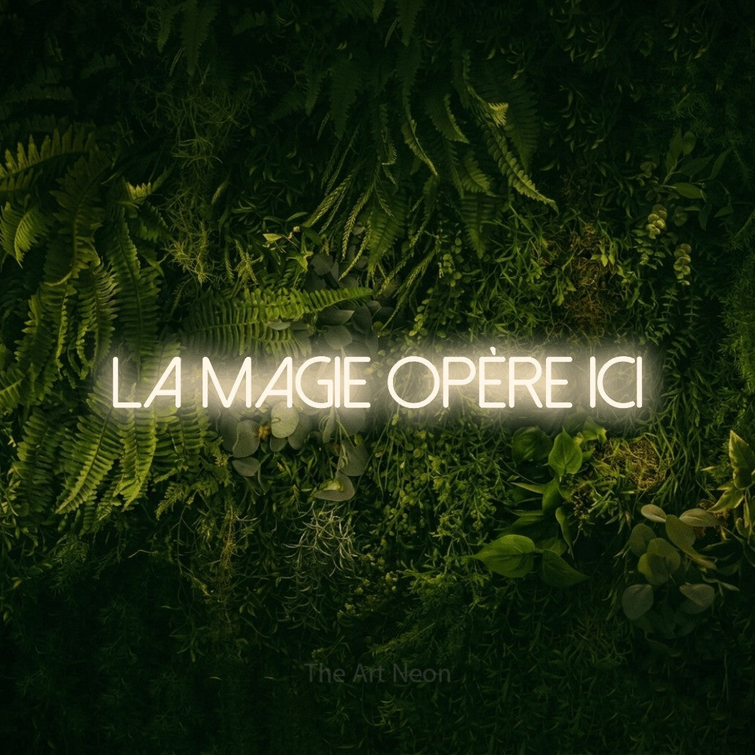 “La magie opère ici” LED neon sign in warm white, with thin sans-serif lettering, shown against a dark moss and forest backdrop.