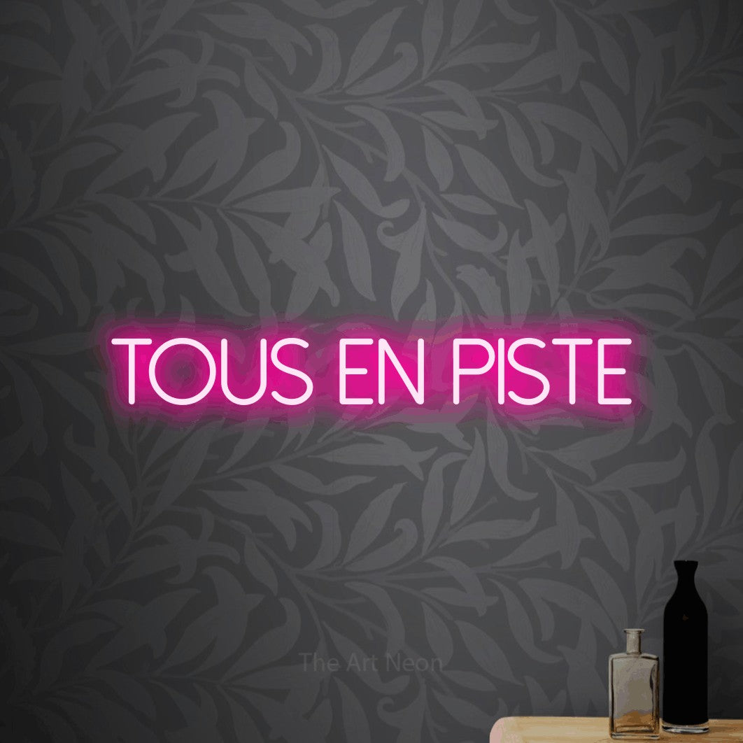 “Tous en piste” LED neon sign in hot pink, with thin sans-serif lettering, shown against grey botanical wallpaper.