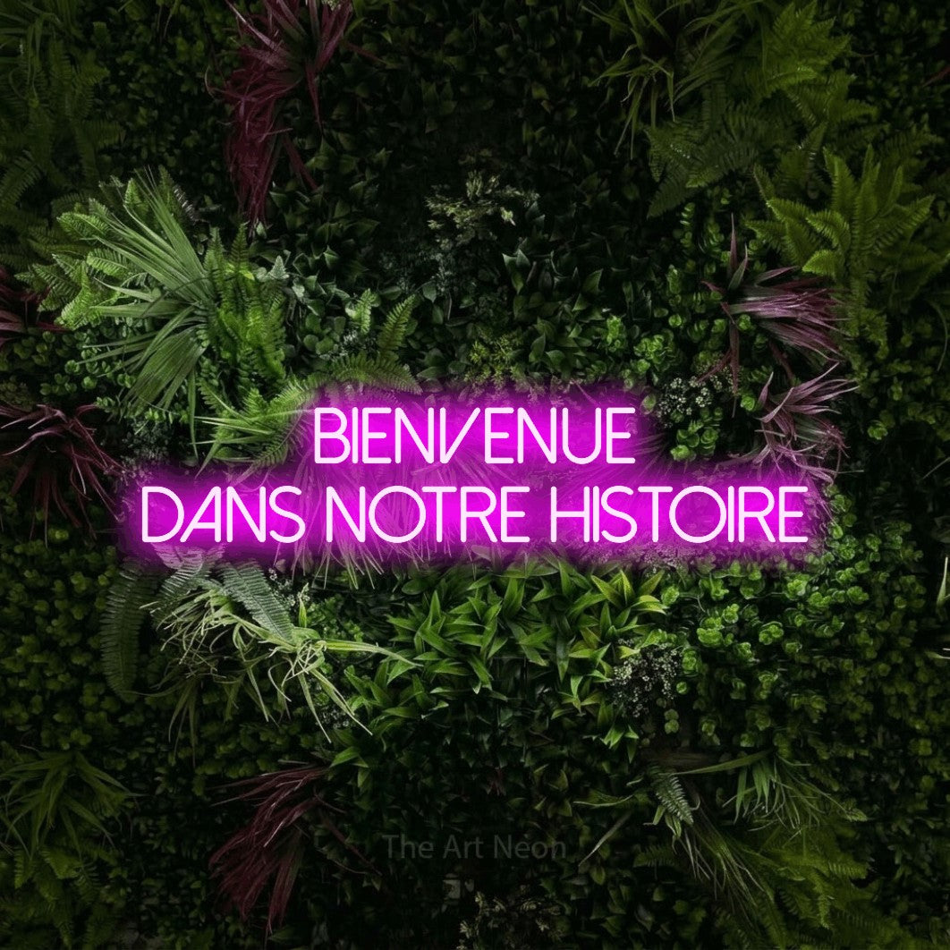 Enseigne néon LED « Bienvenue dans notre histoire » sur deux lignes en rose, en lettres fines sans empattement, présentée sur un mur végétal aux fleurs roses, pour une réception de mariage.