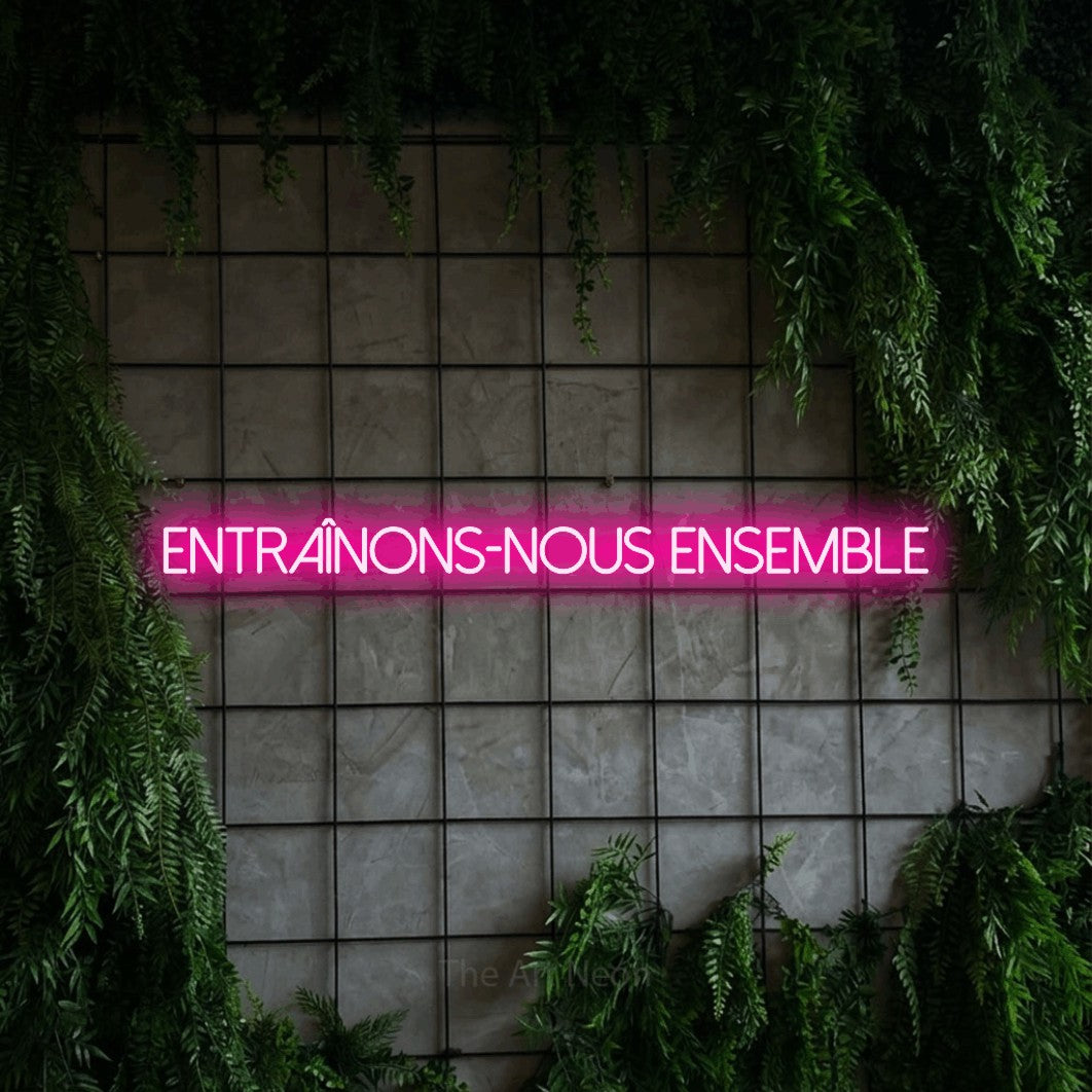 Lettrage en néon LED « Entraînons-nous ensemble » en rose vif, en lettres fines sans empattement, présenté sur une grille de béton couverte de lierre, pour un espace d’entraînement.