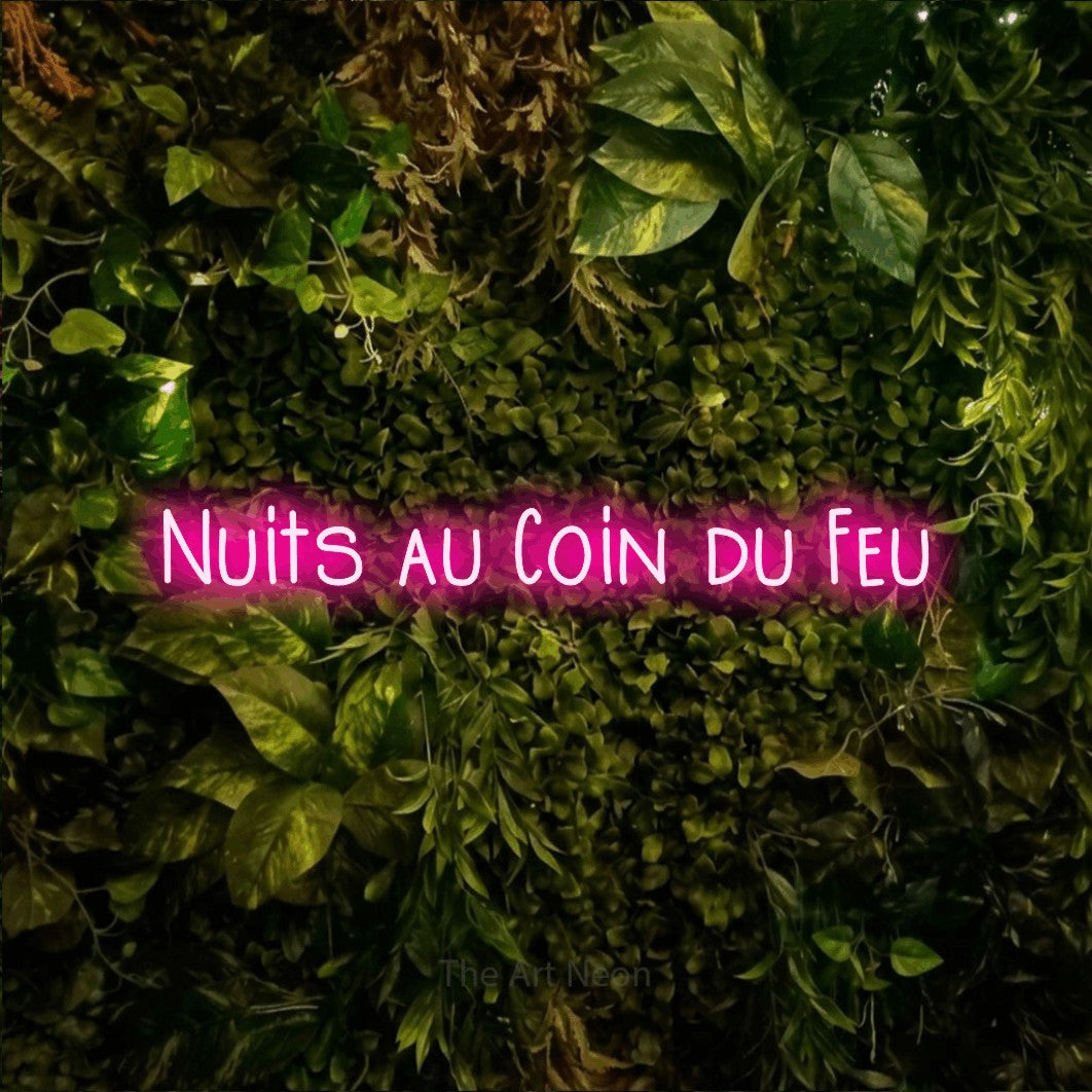 “Nuits au coin du feu” LED neon sign in hot pink, with narrow hand-drawn lettering, shown against bright green and yellow foliage.