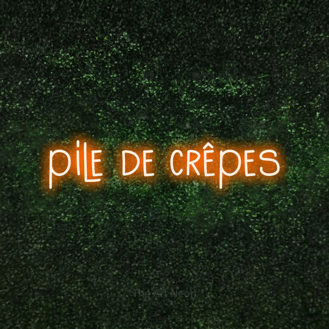 “Pile de crêpes” LED neon sign in orange, with narrow hand-drawn lettering, shown against a textured green hedge.