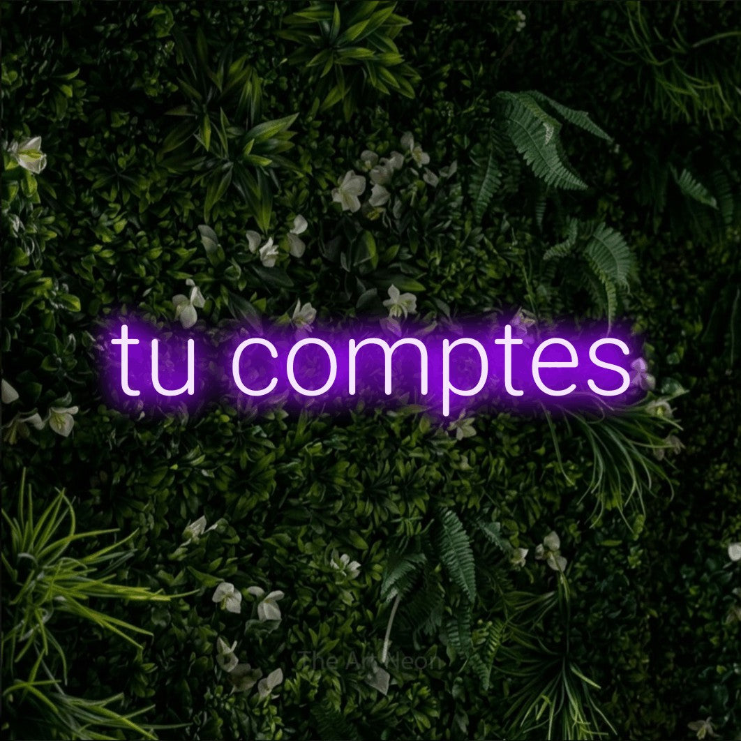 “Tu comptes” LED neon sign in purple, with rounded sans-serif lettering, shown against a dense green plant wall.