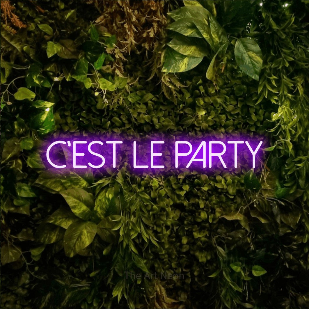 “C'est le party” LED neon sign in purple, with thin sans-serif lettering, shown against bright green and yellow foliage.