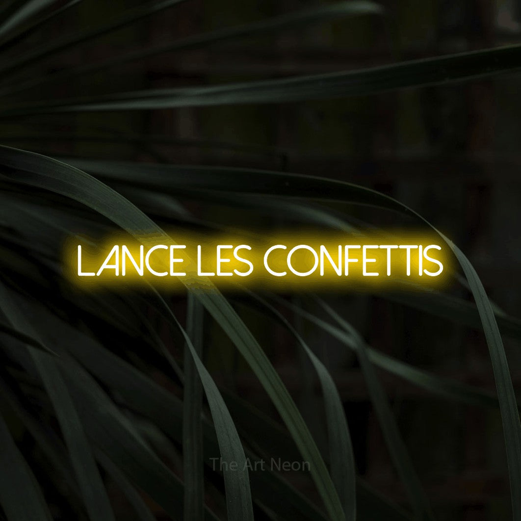 Composition en néon LED « Lance les confettis » en jaune, en lettres fines sans empattement, mise en valeur sur un feuillage sombre en gros plan, pour une fête.