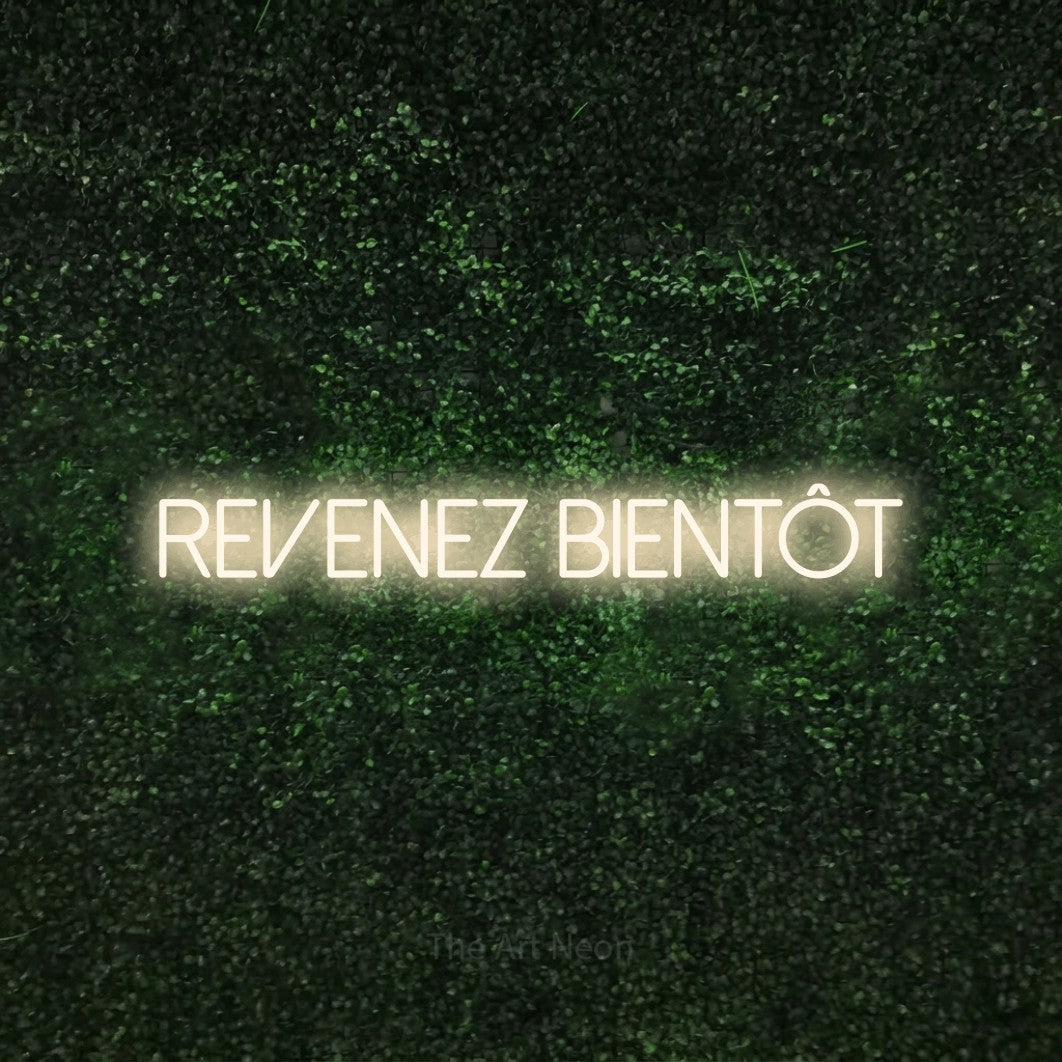 “Revenez bientôt” LED neon sign in warm white, with thin sans-serif lettering, shown against a textured green hedge.