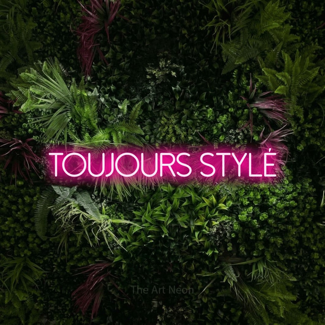 Enseigne néon LED « Toujours stylé » en rose vif, en lettres fines sans empattement, présentée sur un mur végétal aux fleurs roses, pour une boutique ou un commerce.