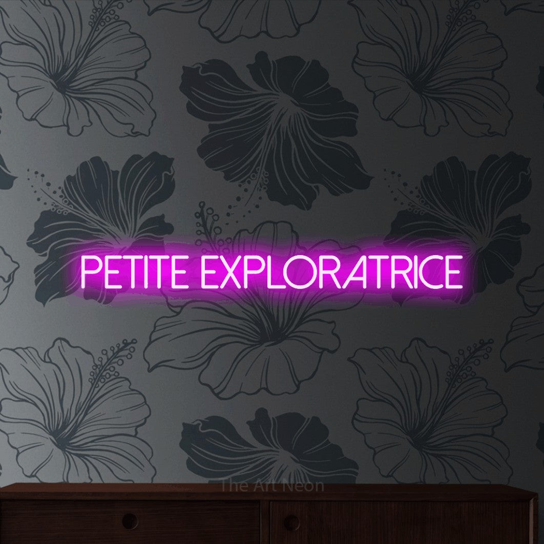 “Petite exploratrice” LED neon sign in pink, with thin sans-serif lettering, shown against a grey floral-patterned wall.