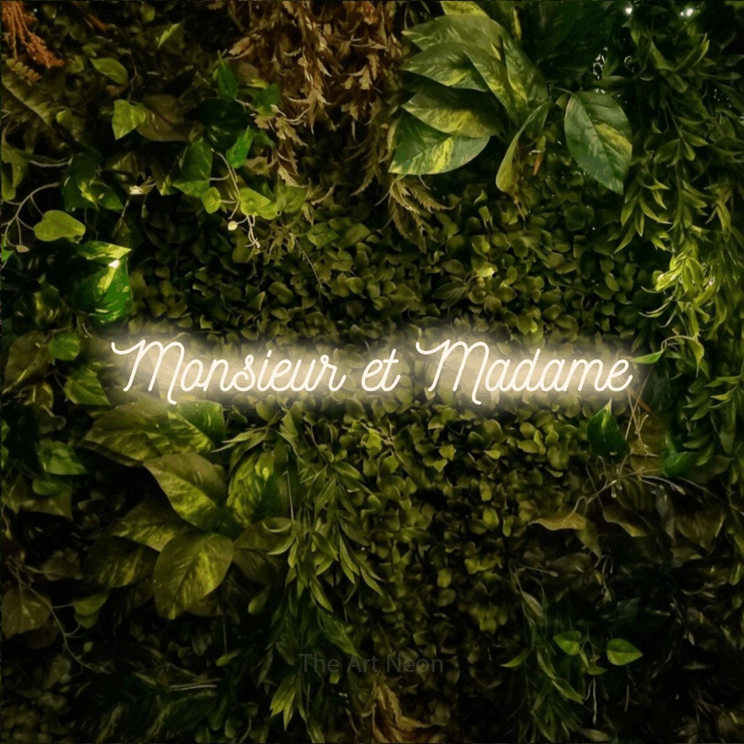 “Monsieur et madame” LED neon sign in warm white, with ornate cursive lettering, shown against bright green and yellow foliage.