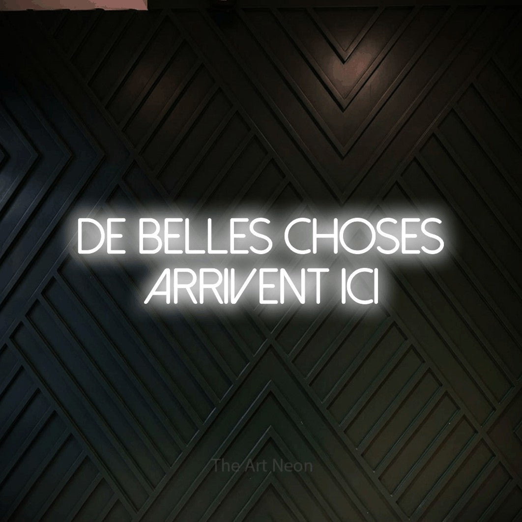 “De belles choses arrivent ici” LED neon sign in cool white, with thin sans-serif lettering across 2 lines, shown against a black chevron-patterned wood wall.