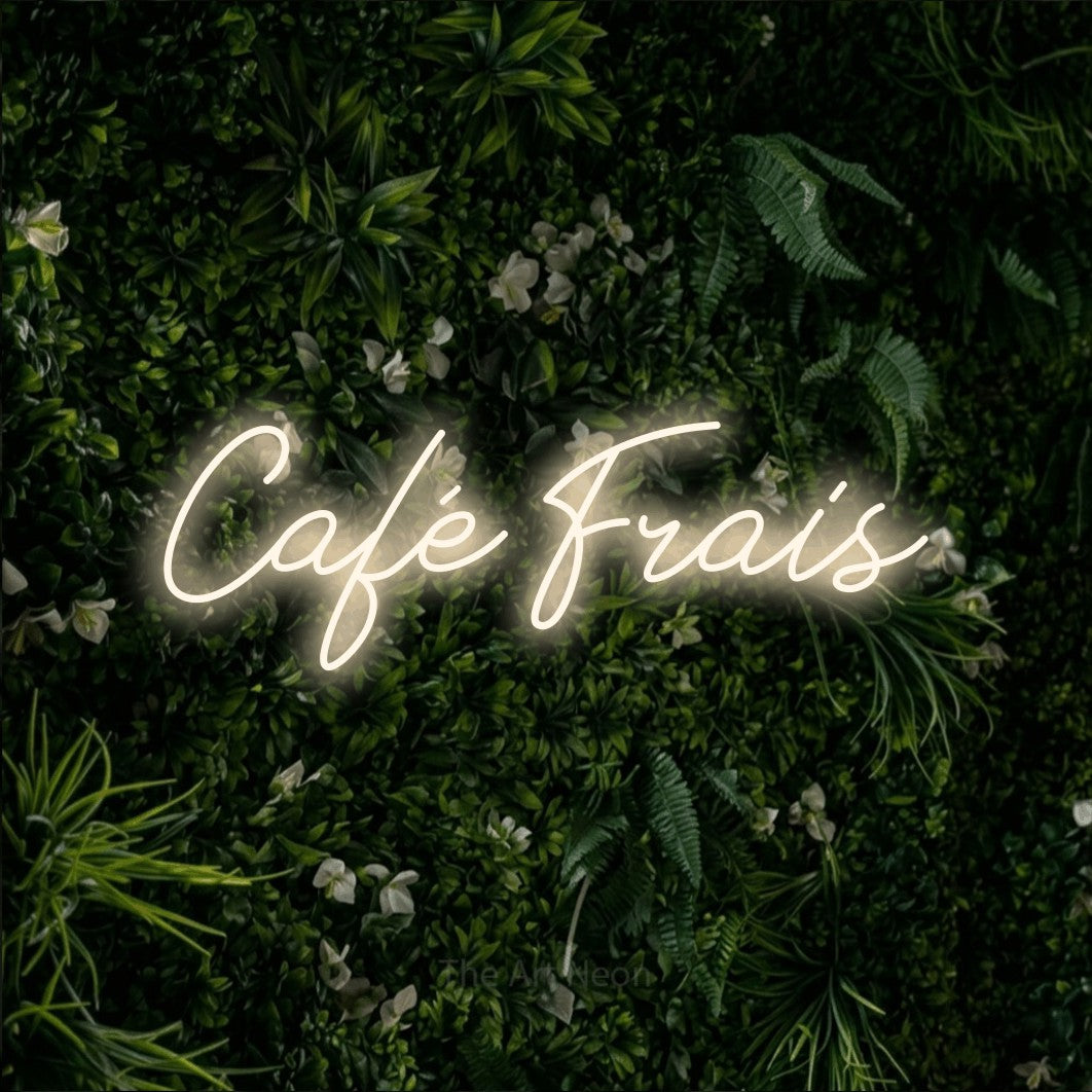“Café frais” LED neon sign in warm white, with cursive lettering, shown against a dense green plant wall.