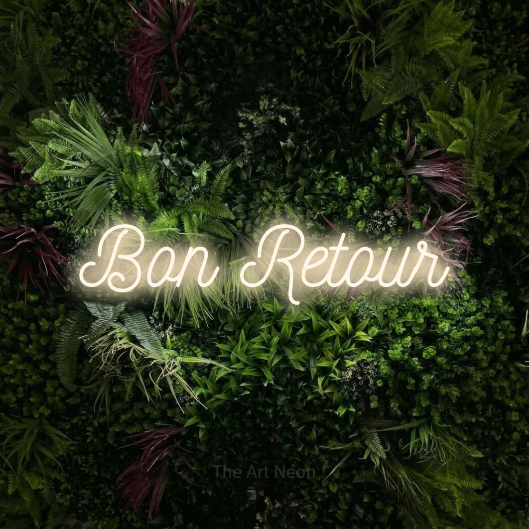 “Bon retour” LED neon sign in warm white, with ornate cursive lettering, shown against a plant wall with pink flowers.