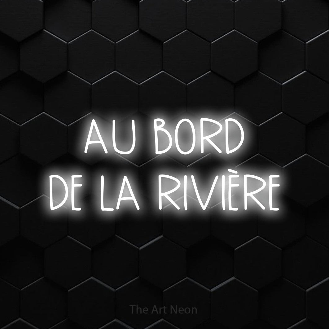 “Au bord de la rivière” LED neon sign in cool white, with narrow hand-drawn lettering across 2 lines, shown against black hexagonal wall tiles.