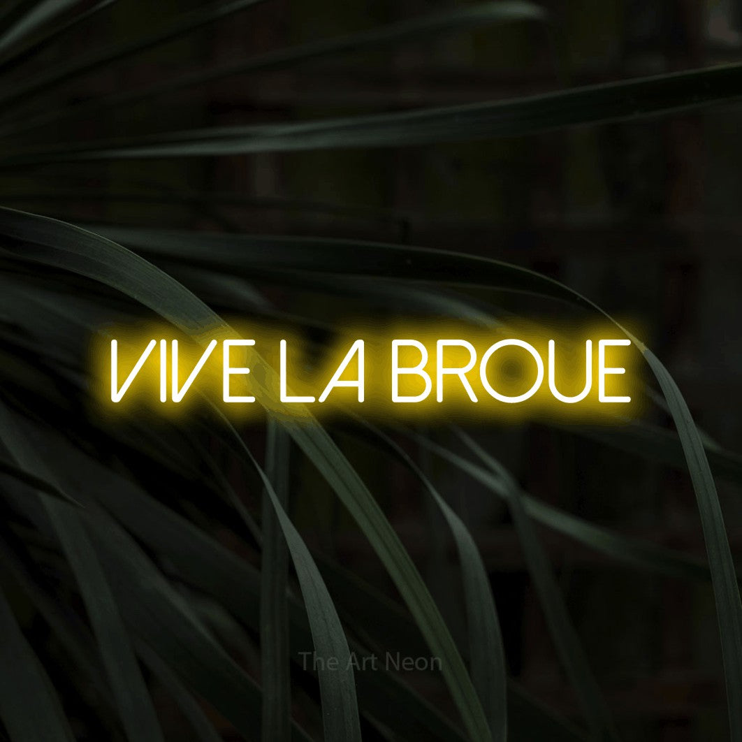 “Vive la broue” LED neon sign in yellow, with thin sans-serif lettering, shown against a close-up of dark foliage.