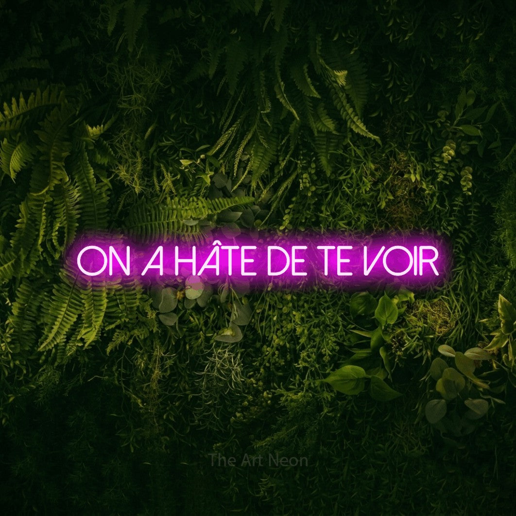 “On a hâte de te voir” LED neon sign in pink, with thin sans-serif lettering, shown against a dark moss and forest backdrop.