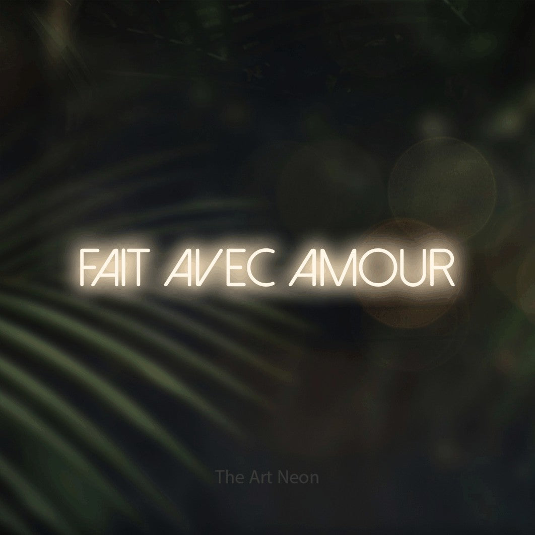 “Fait avec amour” LED neon sign in warm white, with thin sans-serif lettering, shown against a blurred palm and bokeh-light backdrop.
