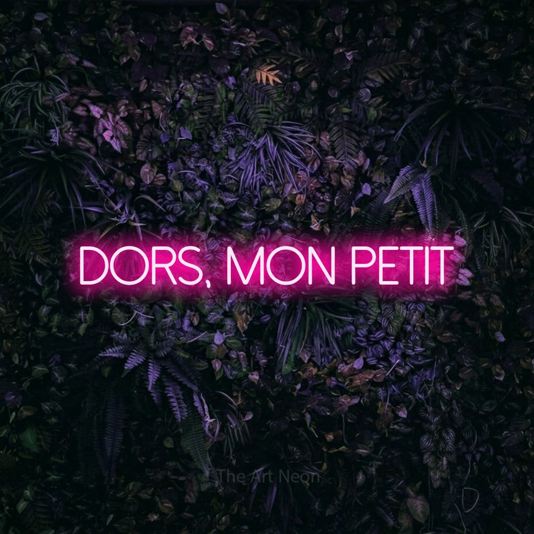 “Dors, mon petit” LED neon sign in hot pink, with thin sans-serif lettering, shown against a dark wall with purple flowers.