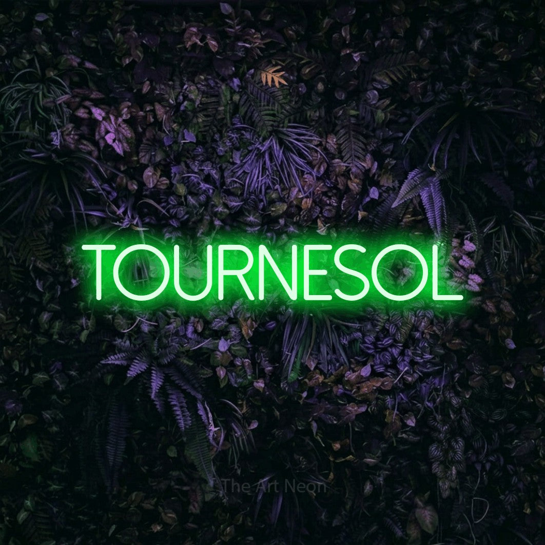 “Tournesol” LED neon sign in green, with thin sans-serif lettering, shown against a dark wall with purple flowers.