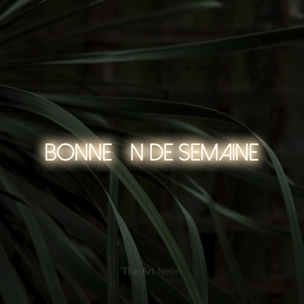 “Bonne fin de semaine” LED neon sign in warm white, with thin sans-serif lettering, shown against a close-up of dark foliage.