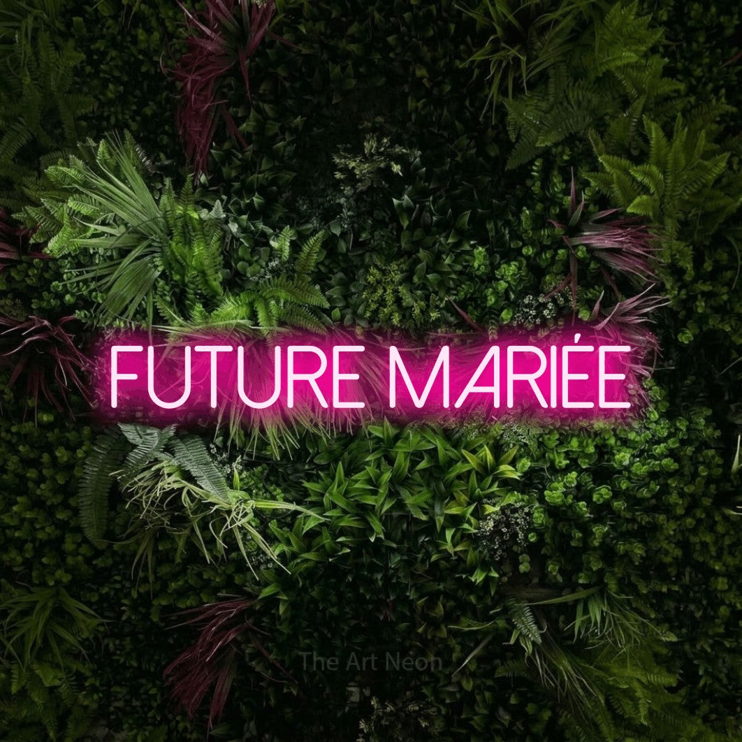 “Future mariée” LED neon sign in hot pink, with thin sans-serif lettering, shown against a plant wall with pink flowers.