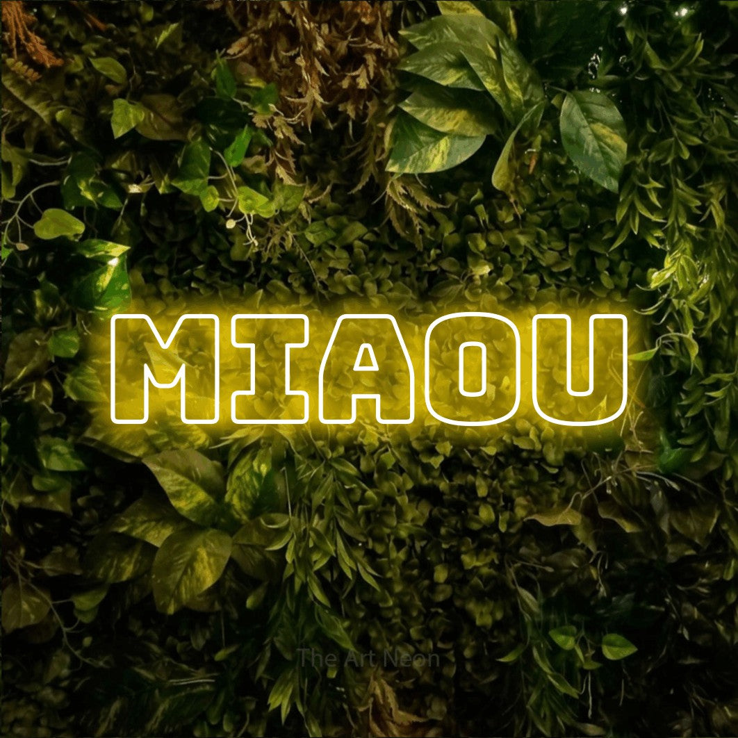 “Miaou” LED neon sign in yellow, with bold outline lettering, shown against bright green and yellow foliage.