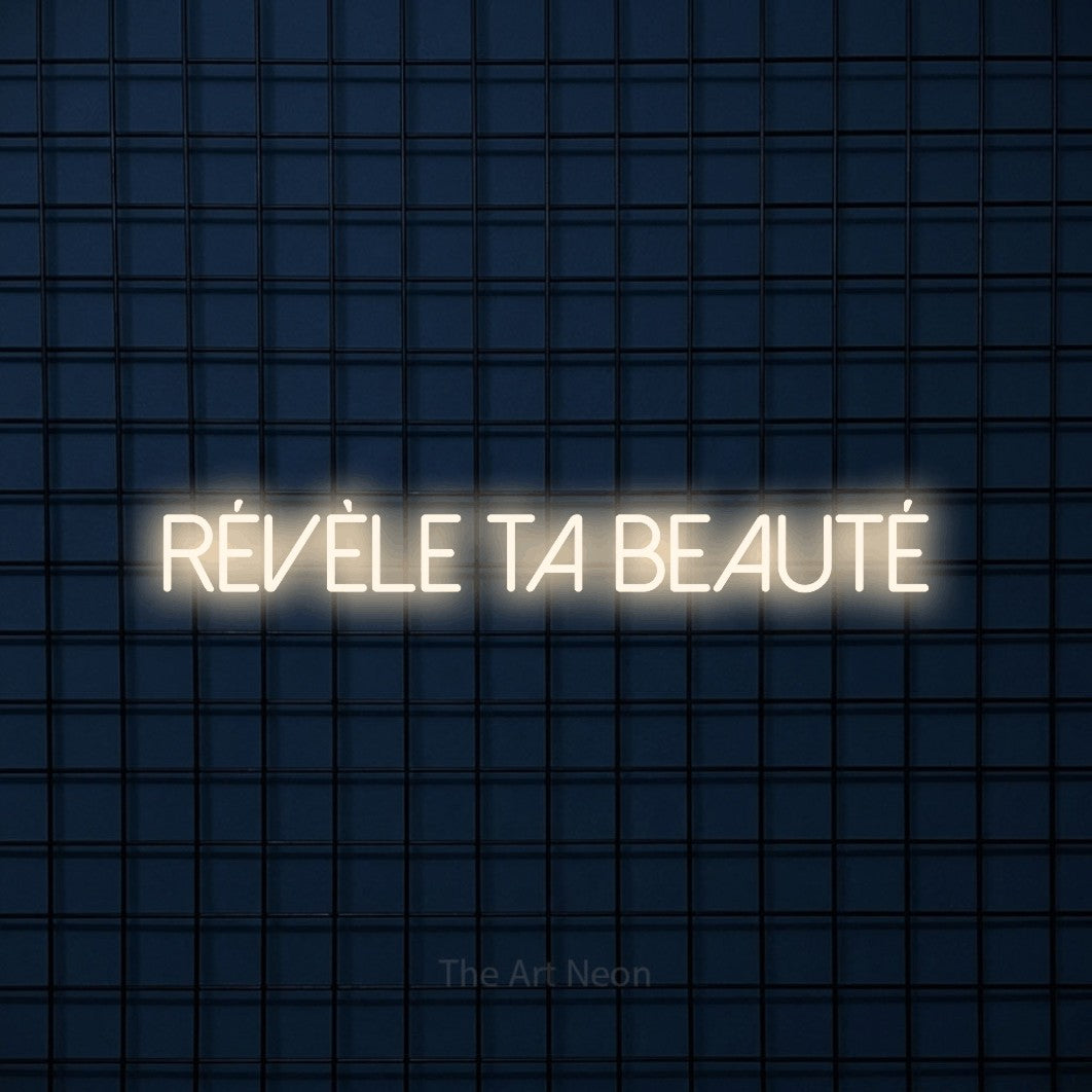 Décoration murale en néon LED « Révèle ta beauté » en blanc chaud, en lettres fines sans empattement, installée sur une grille bleu foncé, pour un salon de beauté.