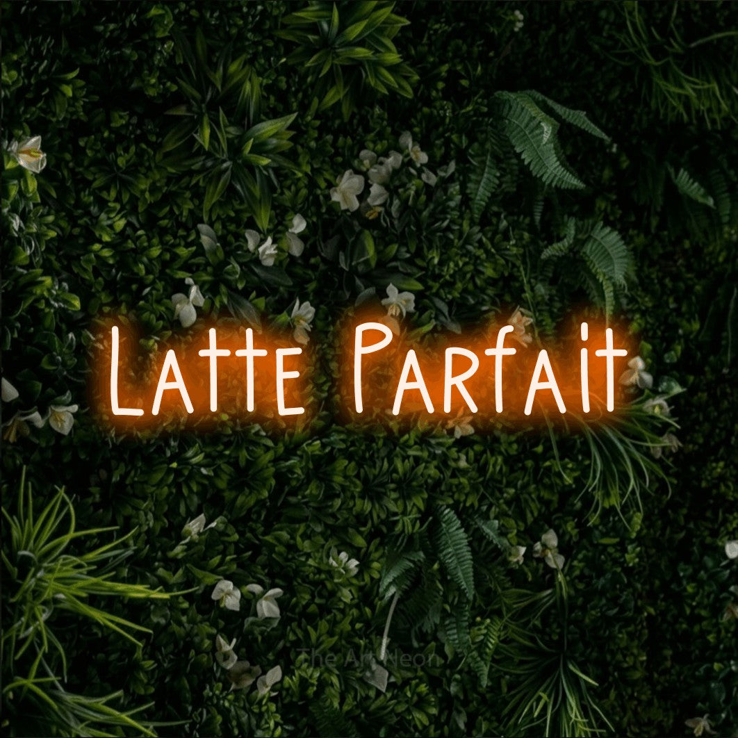 “Latte parfait” LED neon sign in orange, with narrow hand-drawn lettering, shown against a dense green plant wall.