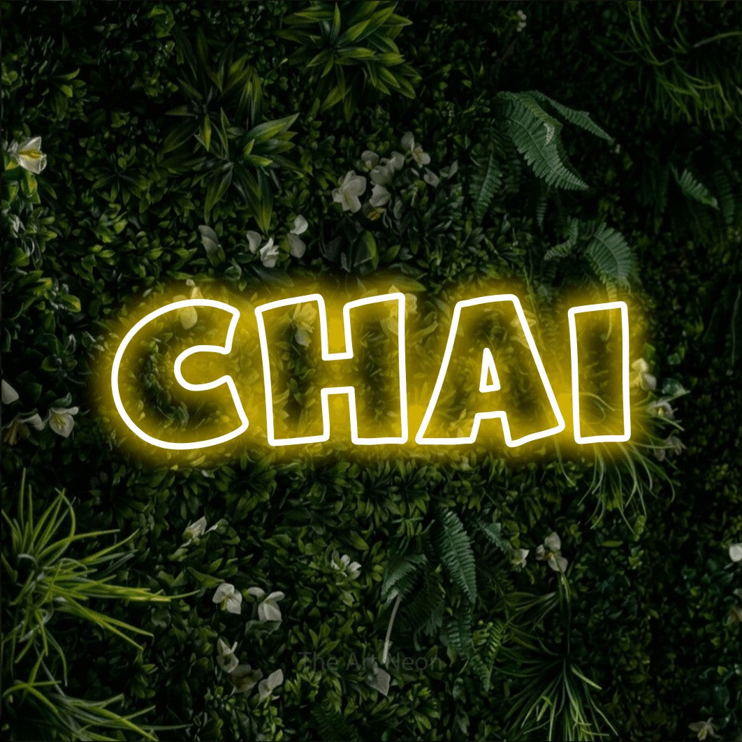 “Chai” LED neon sign in yellow, with wide lettering with irregular outlines, shown against a dense green plant wall.