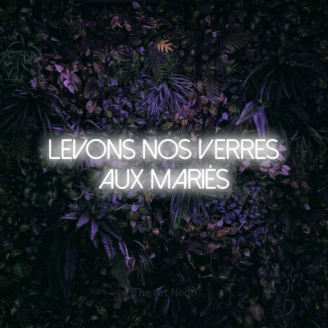 “Levons nos verres aux mariés” LED neon sign in cool white, with thin sans-serif lettering across 2 lines, shown against a dark wall with purple flowers.