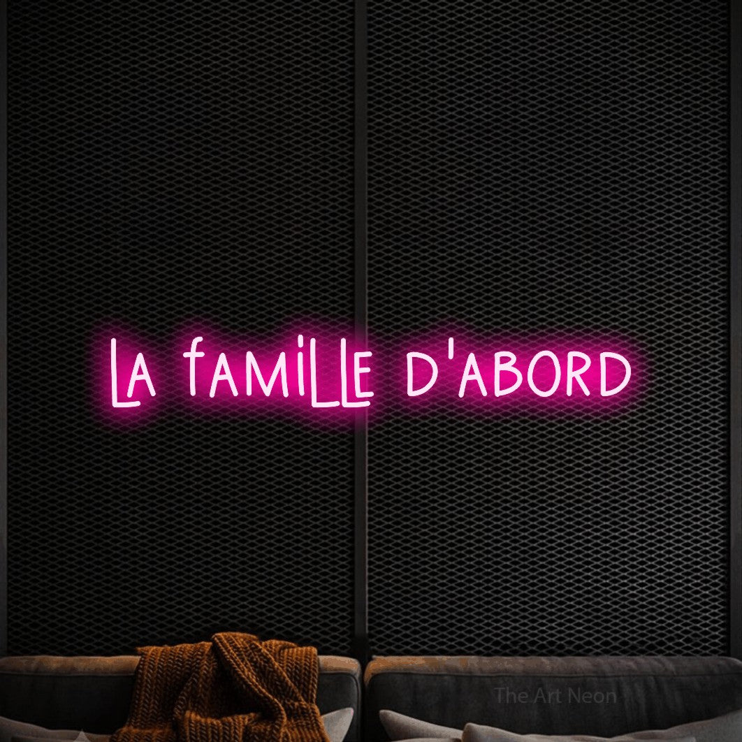 “La famille d'abord” LED neon sign in hot pink, with narrow hand-drawn lettering, shown against a modern dark-toned seating area.