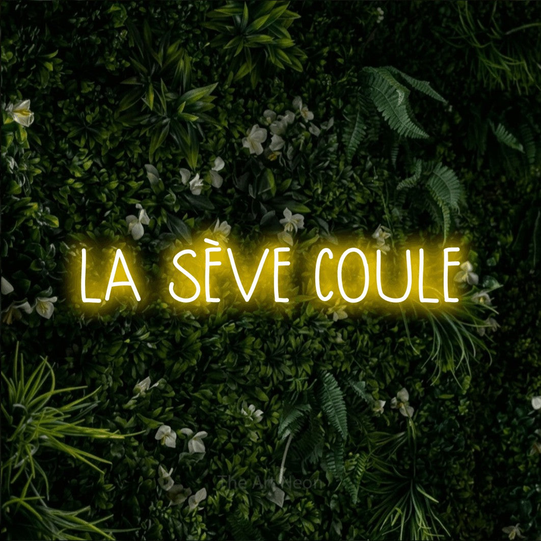 “La sève coule” LED neon sign in yellow, with narrow hand-drawn lettering, shown against a dense green plant wall.