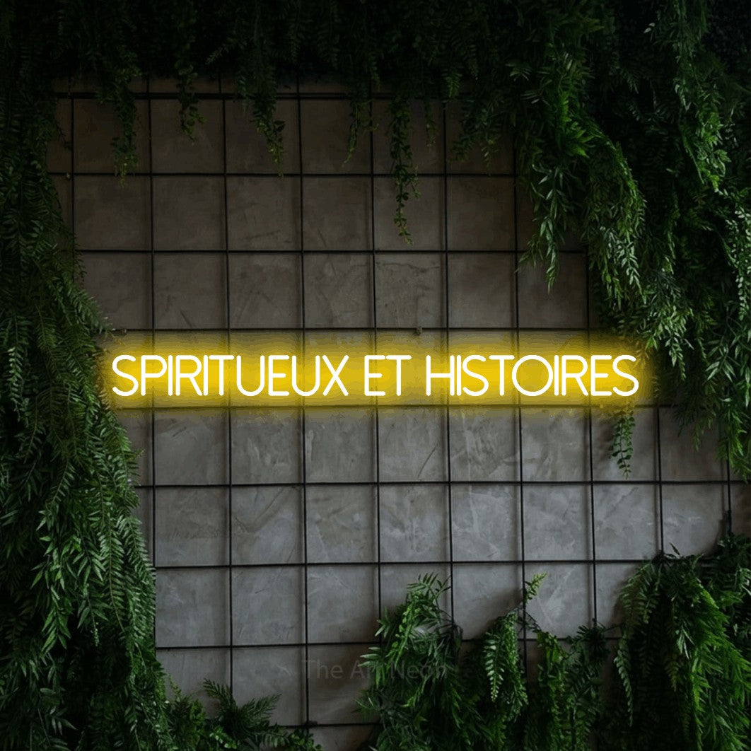 “Spiritueux et histoires” LED neon sign in yellow, with thin sans-serif lettering, shown against an ivy-covered concrete grid.