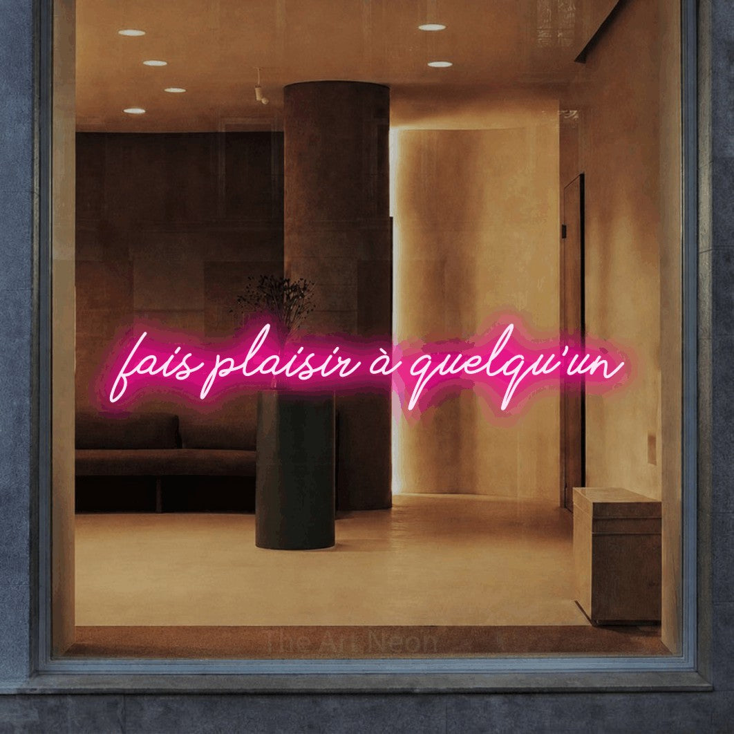 “Fais plaisir à quelqu'un” LED neon sign in hot pink, with cursive lettering, shown against a warm-toned interior.