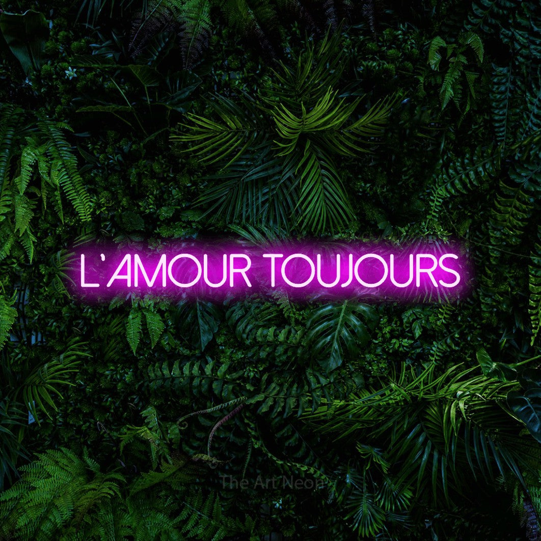 “L'amour toujours” LED neon sign in pink, with thin sans-serif lettering, shown against dense tropical foliage.