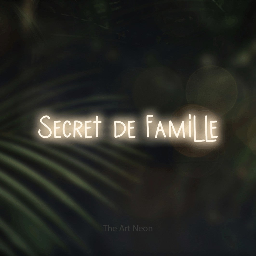 “Secret de famille” LED neon sign in warm white, with narrow hand-drawn lettering, shown against a blurred palm and bokeh-light backdrop.