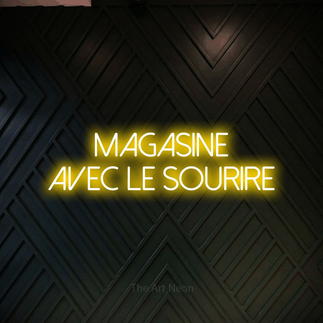 “Magasine avec le sourire” LED neon sign in yellow, with thin sans-serif lettering across 2 lines, shown against a black chevron-patterned wood wall.