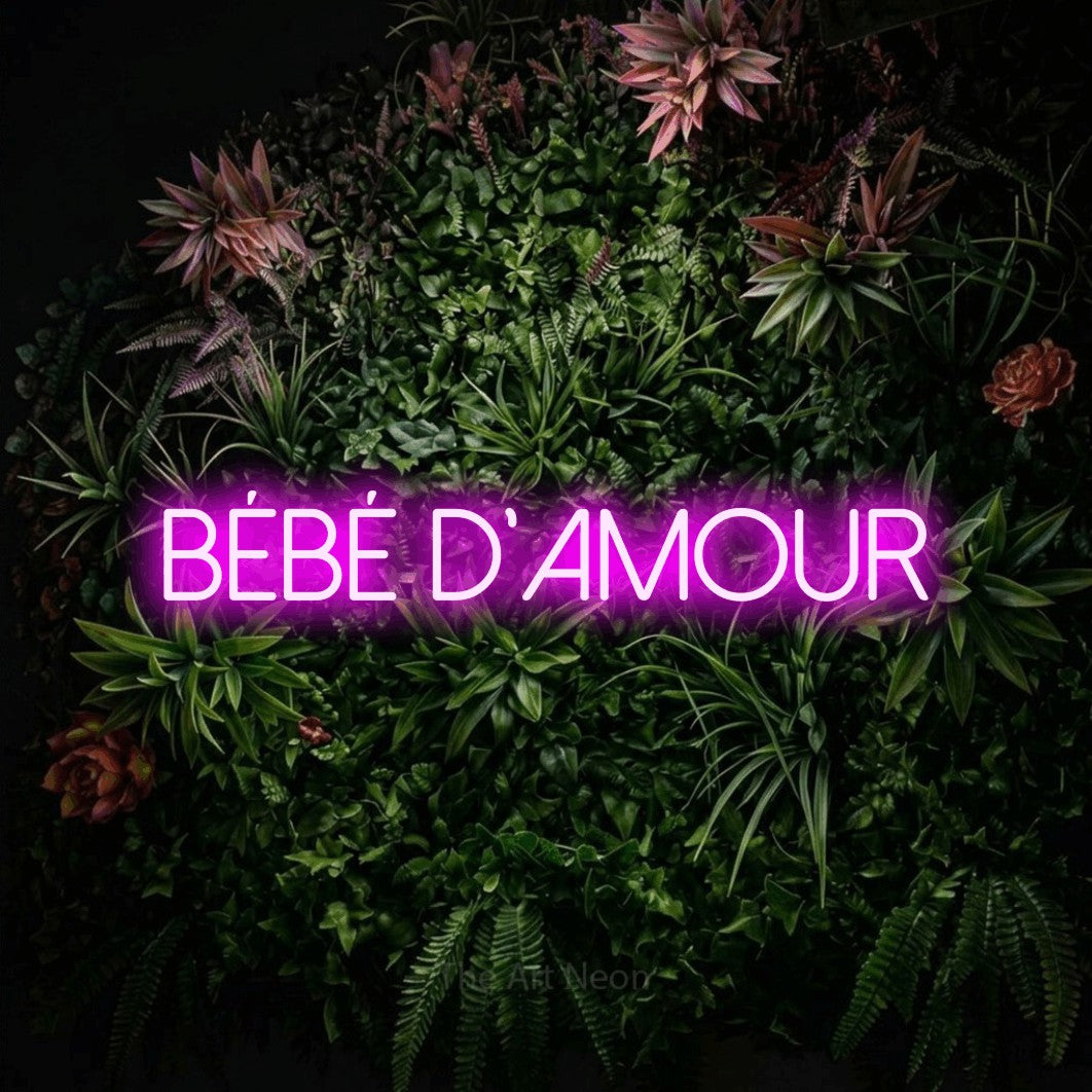 “Bébé d'amour” LED neon sign in pink, with thin sans-serif lettering, shown against a flowering plant wall.