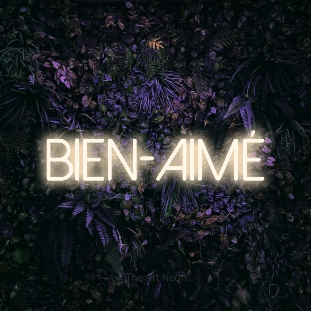 “Bien-aimé” LED neon sign in warm white, with thin sans-serif lettering, shown against a dark wall with purple flowers.