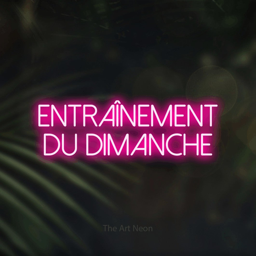 “Entraînement du dimanche” LED neon sign in hot pink, with thin sans-serif lettering across 2 lines, shown against a blurred palm and bokeh-light backdrop.