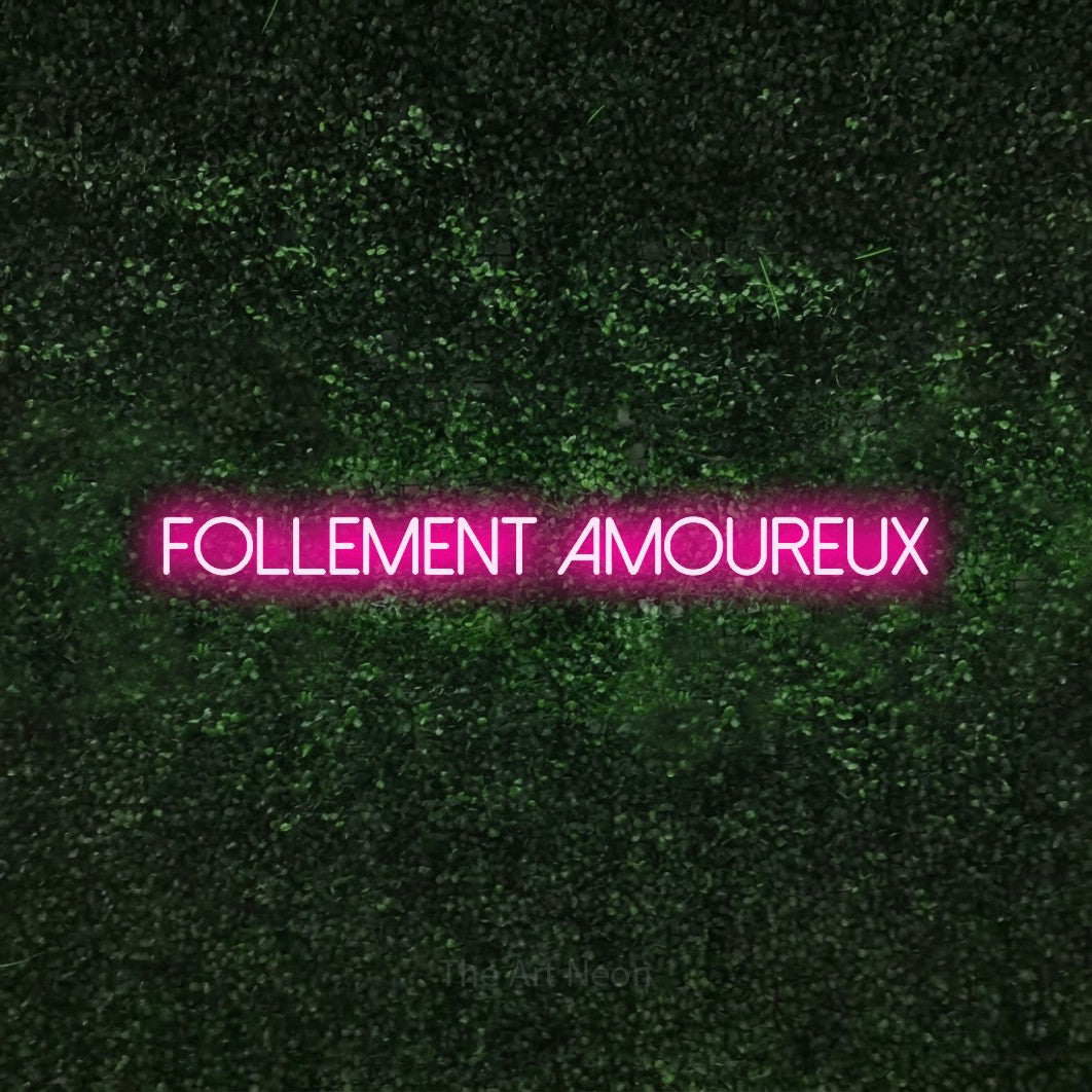 “Follement amoureux” LED neon sign in hot pink, with thin sans-serif lettering, shown against a textured green hedge.