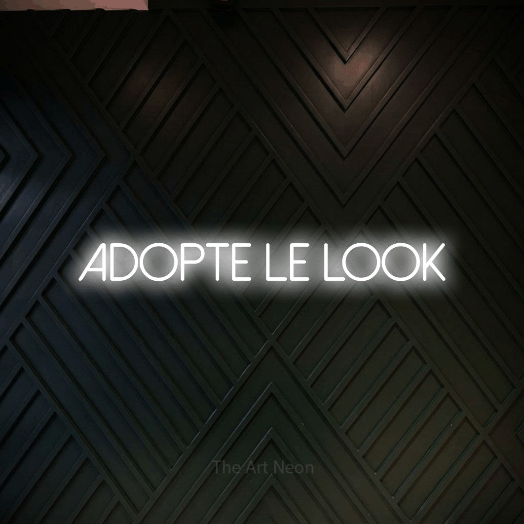 “Adopte le look” LED neon sign in cool white, with thin sans-serif lettering, shown against a black chevron-patterned wood wall.