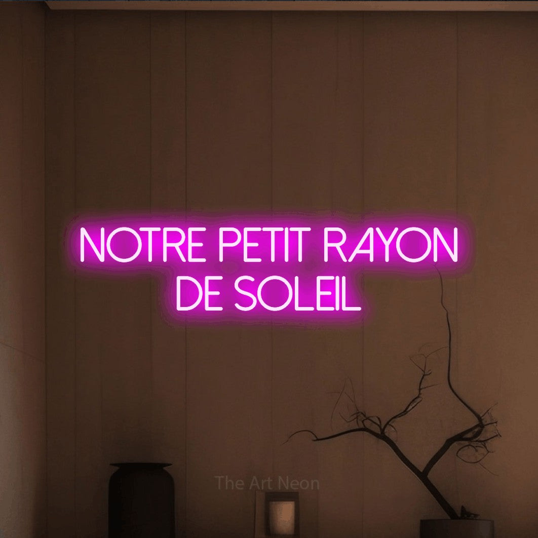 “Notre petit rayon de soleil” LED neon sign in pink, with thin sans-serif lettering across 2 lines, shown against a simple earth-toned interior.