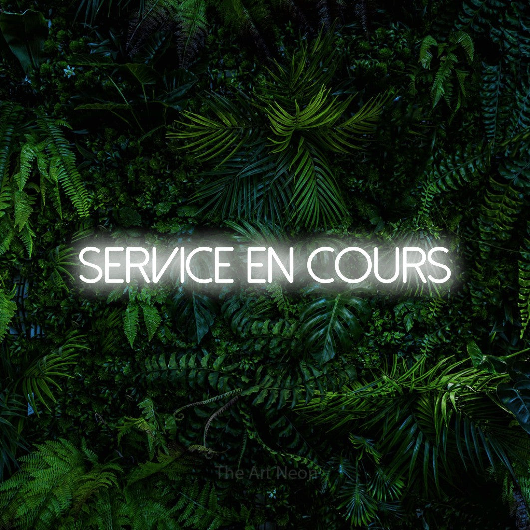 “Service en cours” LED neon sign in cool white, with thin sans-serif lettering, shown against dense tropical foliage.