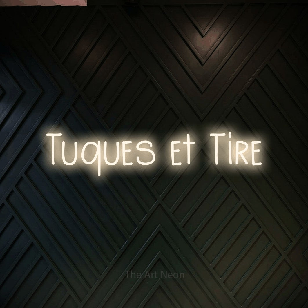 “Tuques et tire” LED neon sign in warm white, with narrow hand-drawn lettering, shown against a black chevron-patterned wood wall.
