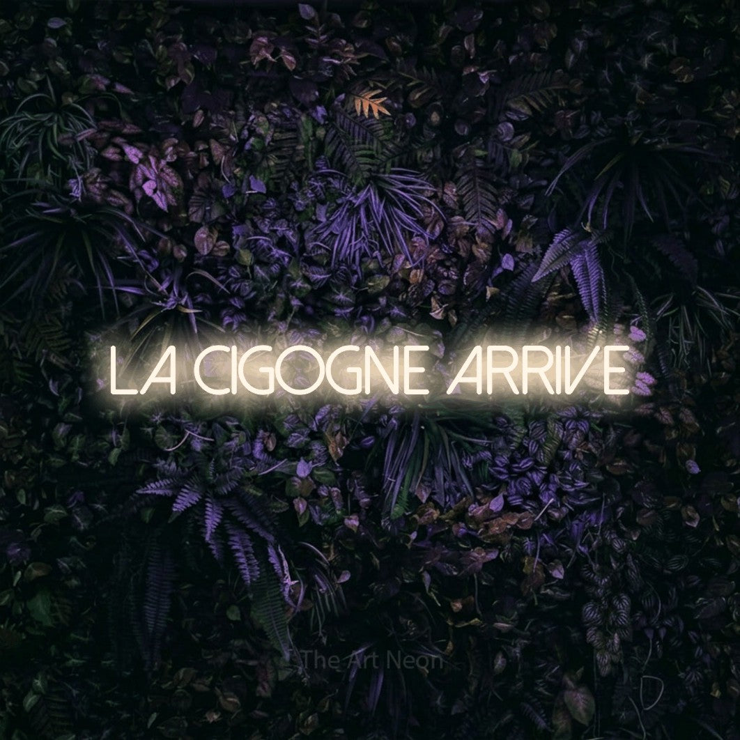 Composition en néon LED « La cigogne arrive » en blanc chaud, en lettres fines sans empattement, mise en valeur sur un mur sombre aux fleurs violettes, pour une fête prénatale.