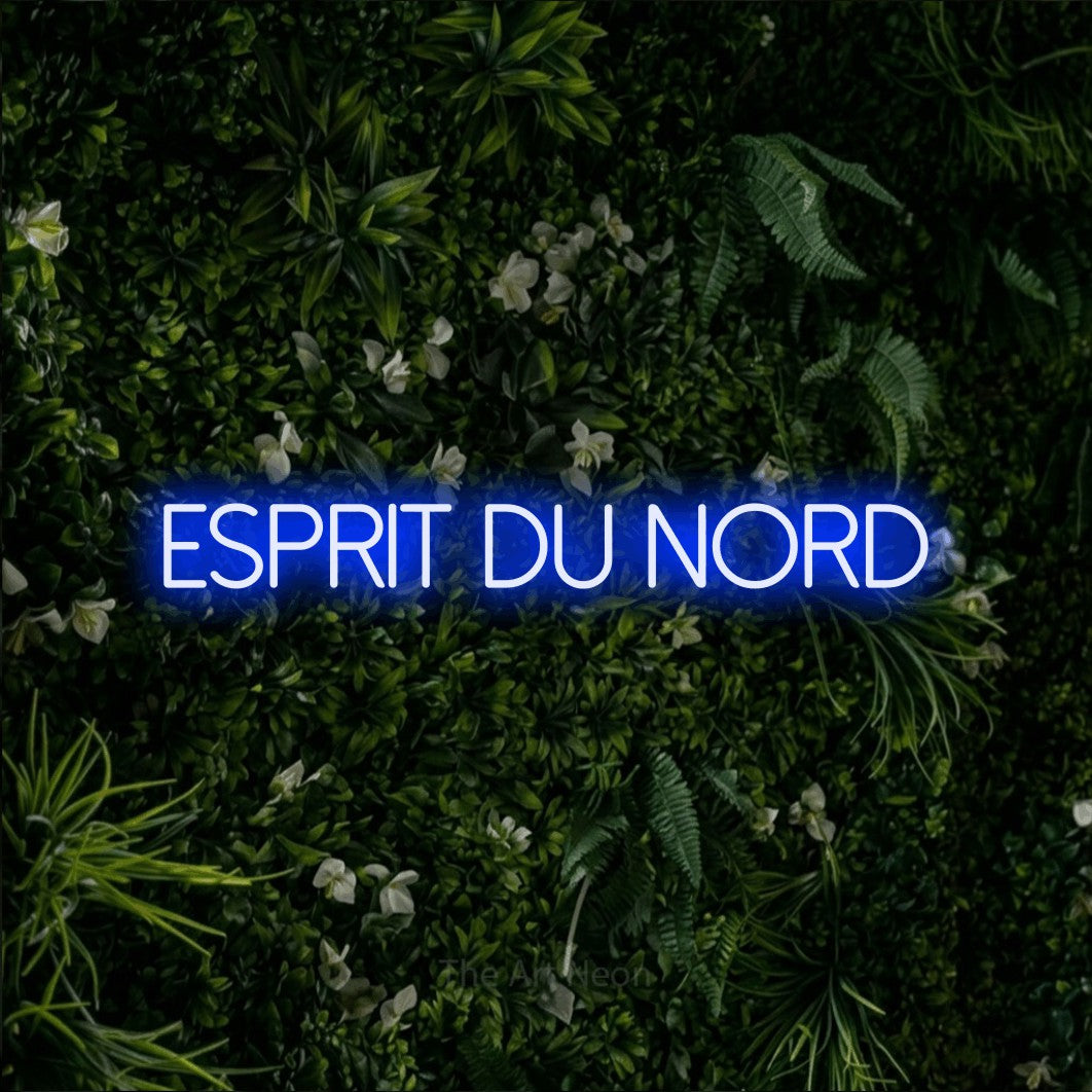 “Esprit du nord” LED neon sign in blue, with thin sans-serif lettering, shown against a dense green plant wall.